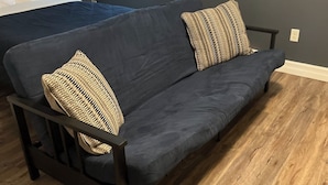 A dark grey sofa with two woven pillows, a small table with a lamp, and a window with blinds. - Stanley House (Clarenville)