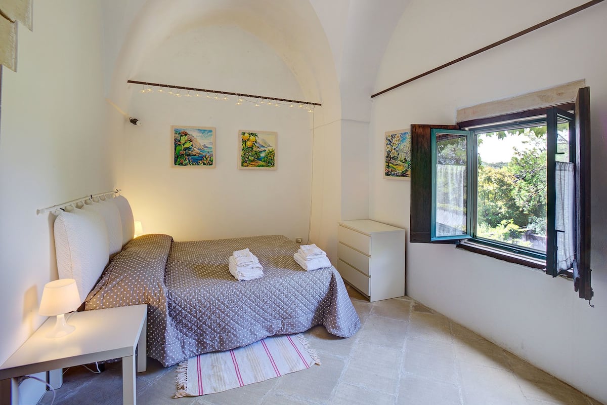 A bedroom with a bed, a bedside table, a nightstand, a window with an open shutter, and wall art.