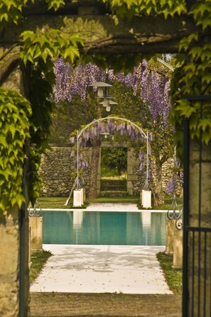 A pool area with a stone wall and a pergola covered in purple flowers. - Tenuta Sant'Andrea (Muro Leccese)