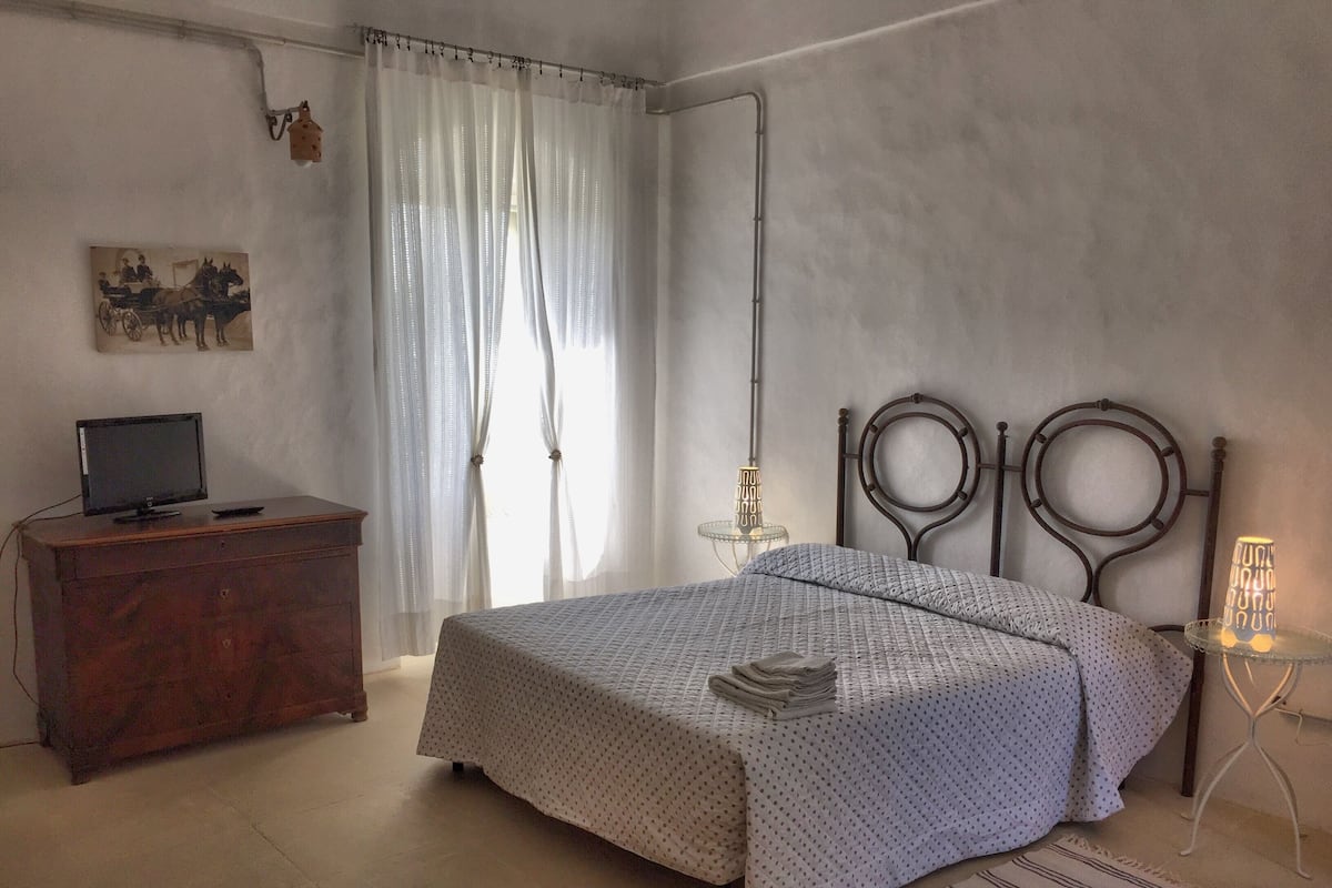 A bedroom with a bed, a wooden dresser, a television on the dresser, a painting on the wall, and a lamp on a side table.