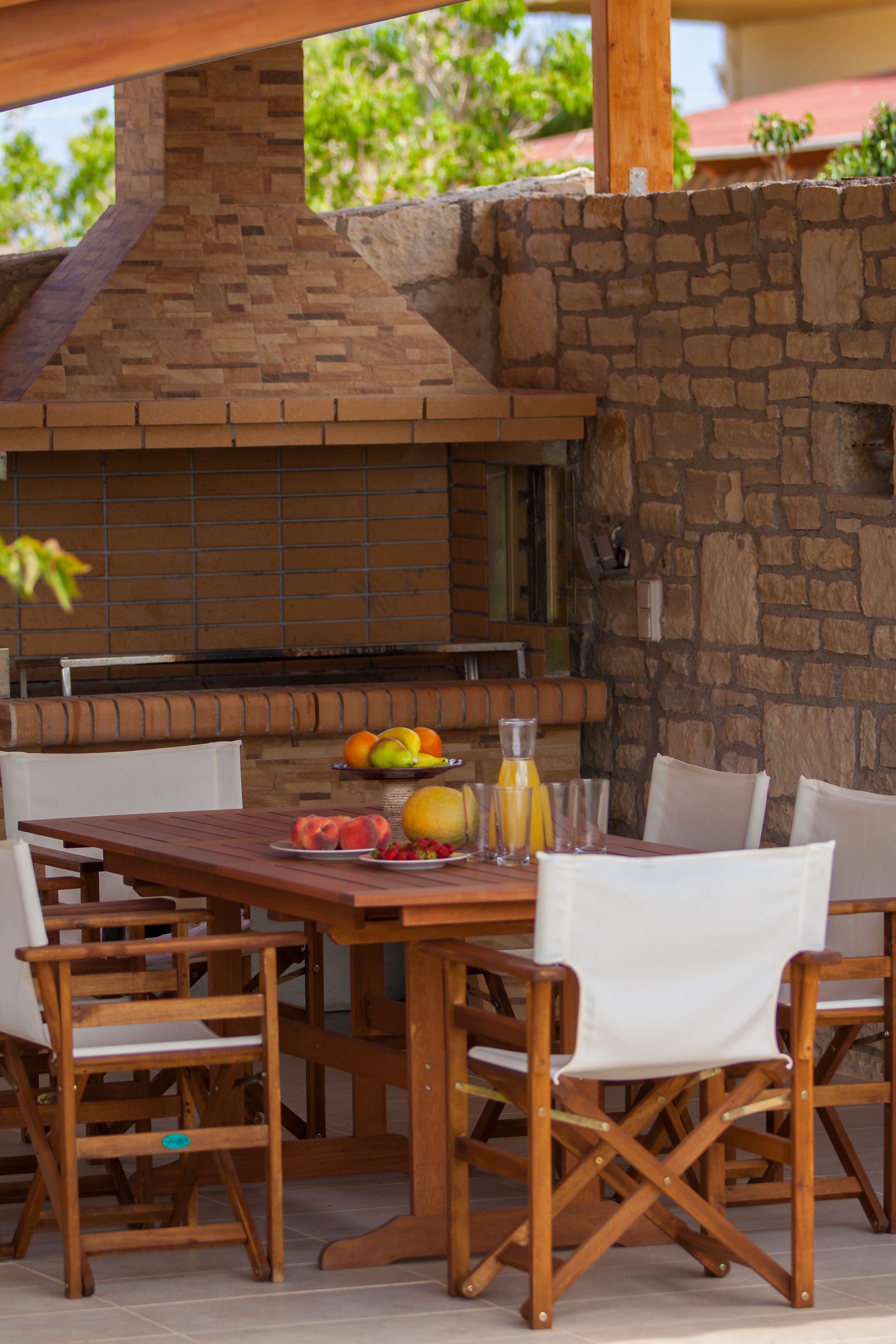 An outdoor dining area with a brick grill, white chairs, and a table set with fruits and a glass of juice.