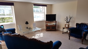 A living room with a blue sofa, a glass coffee table, a television on a wooden stand, and a small side table with a vase. - Apartment 9 Robertson Villas (Rochester)