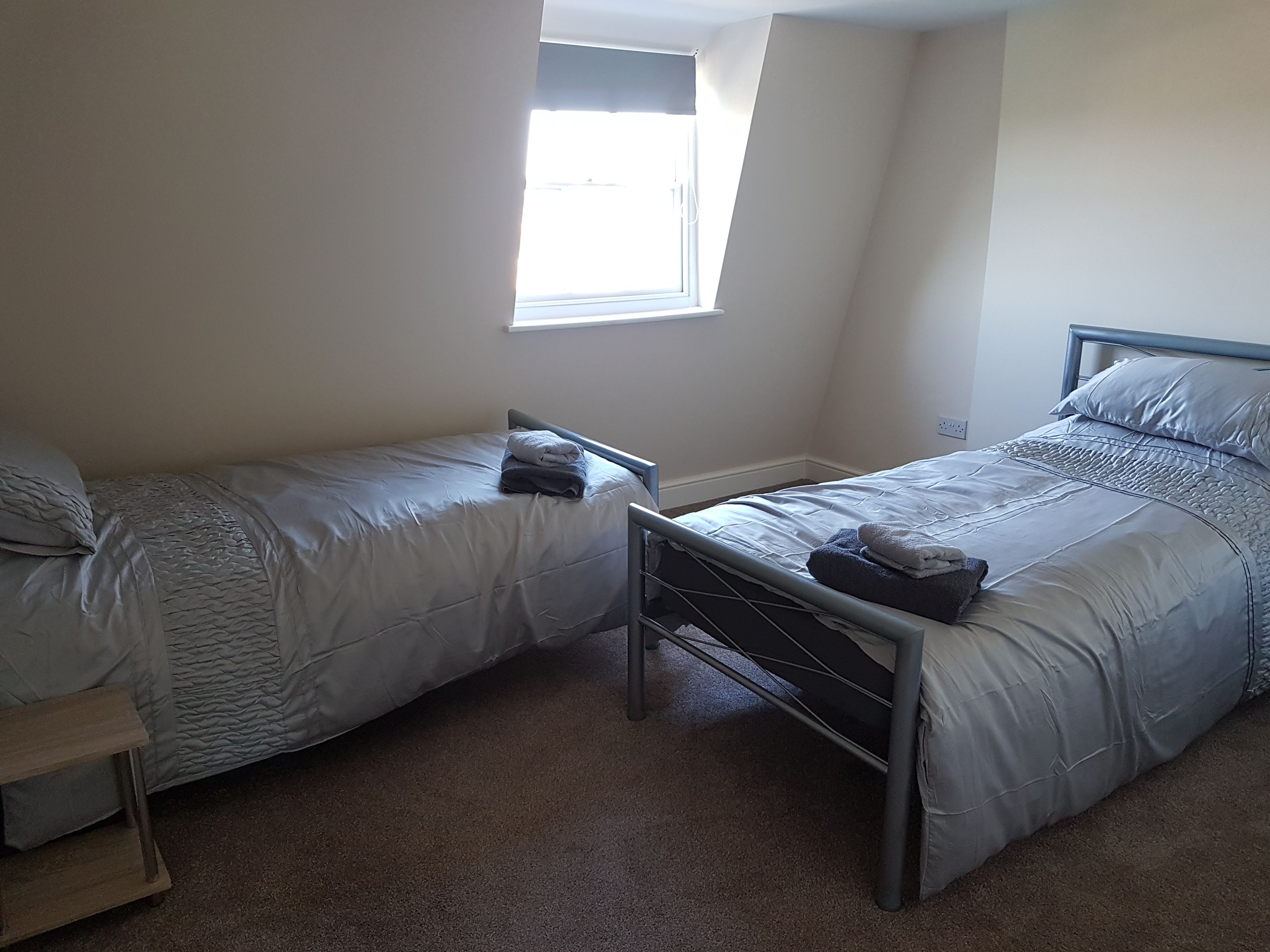 A small, neatly arranged bedroom with two single beds, a window, and a carpeted floor.