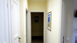 Un couloir aux murs de couleur crème et au plancher en bois, qui mène à une pièce dotée d’une porte et d’un tableau encadré au mur.