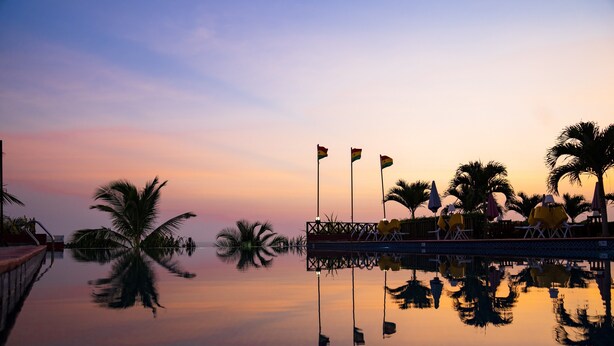 A serene waterfront setting with palm trees and lounge chairs reflecting in calm water, under a sunset sky.