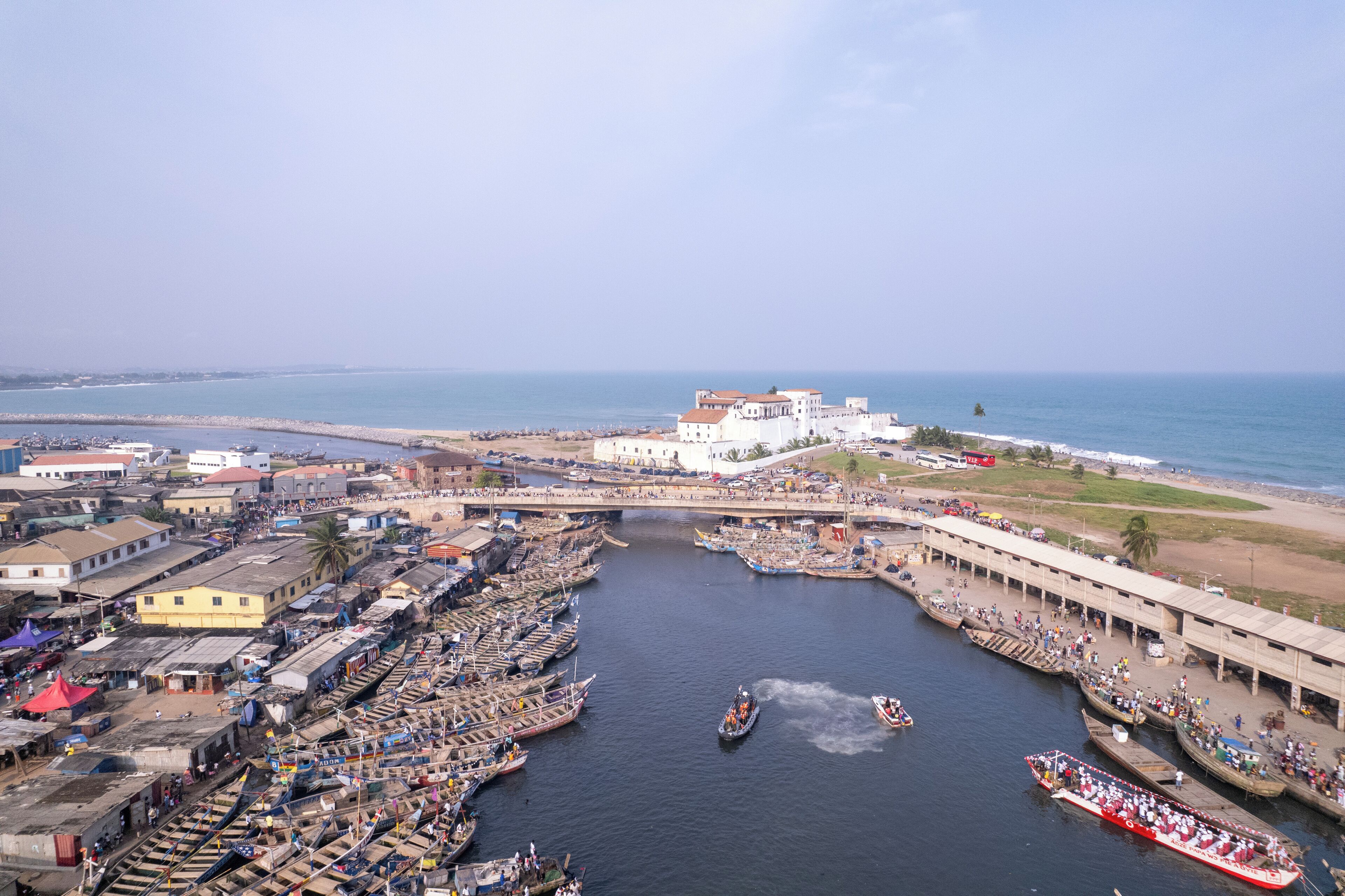 A riverside town with boats docked along the riverbank, a bridge connecting the river to the mainland, and a large building with a red roof in the background.