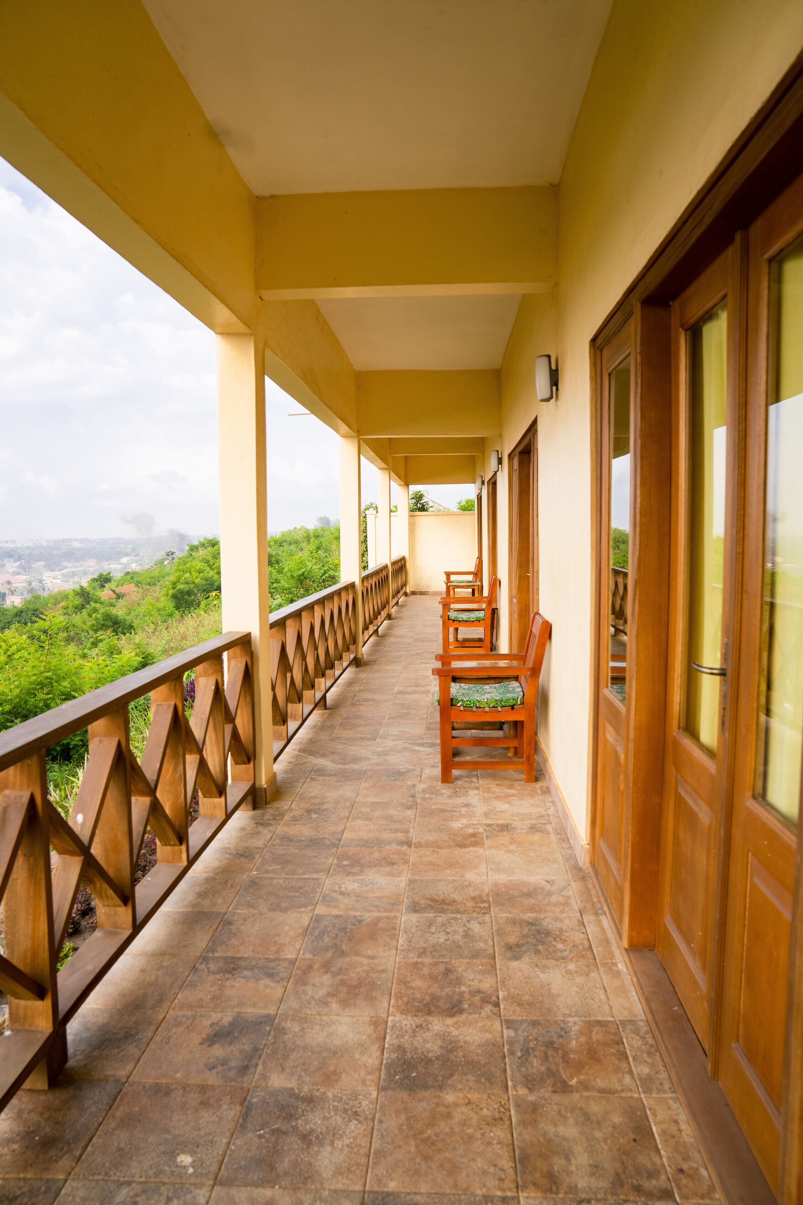 A covered balcony with wooden railings, a tiled floor, and a row of wooden chairs.