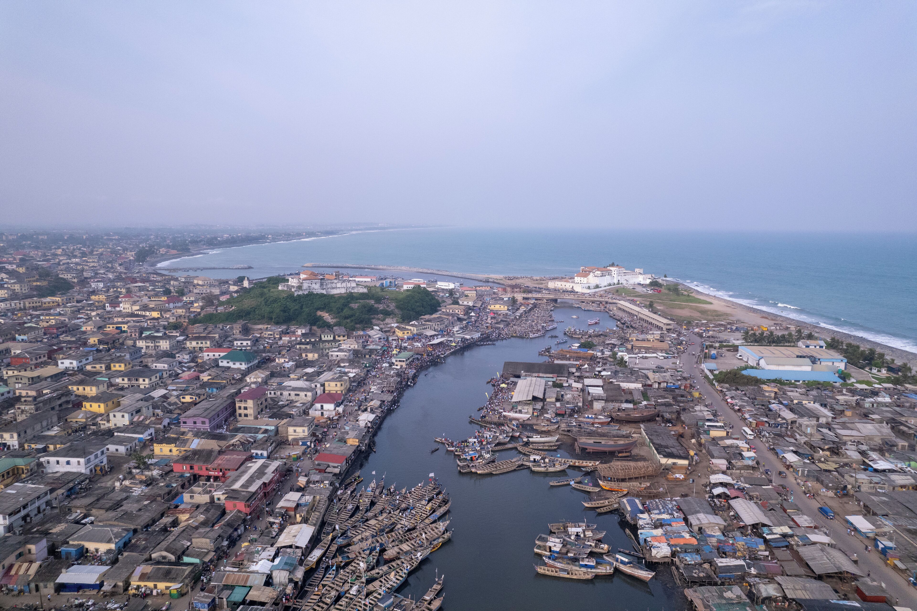 An aerial view of a coastal city with dense urban development, a river running through the center, and a large body of water to the east.