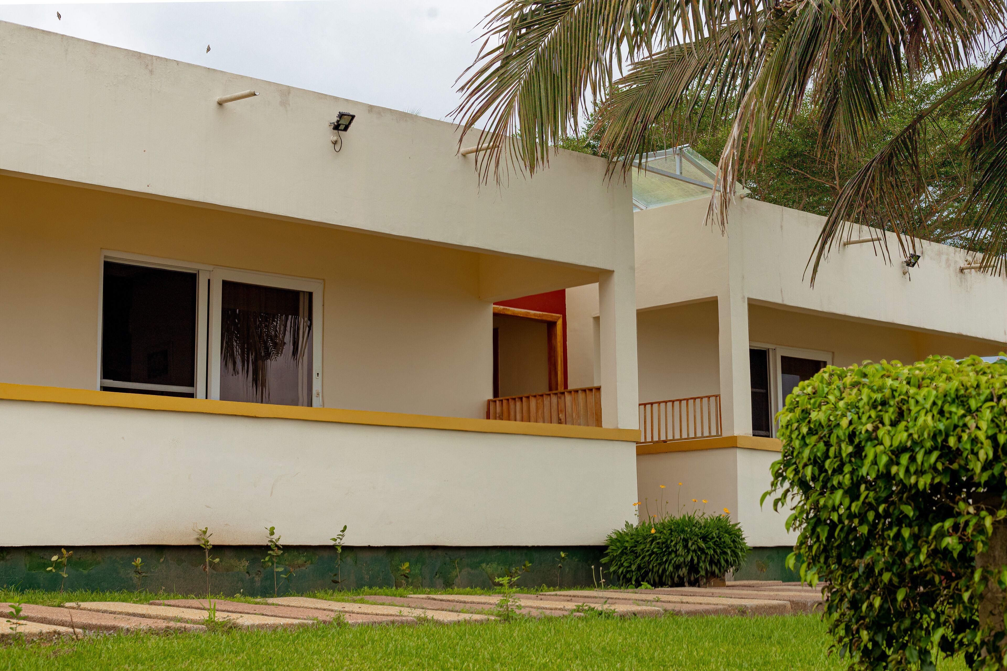 A two-story building with a flat roof, a balcony on the upper floor, and a small garden area with a palm tree and shrubs.