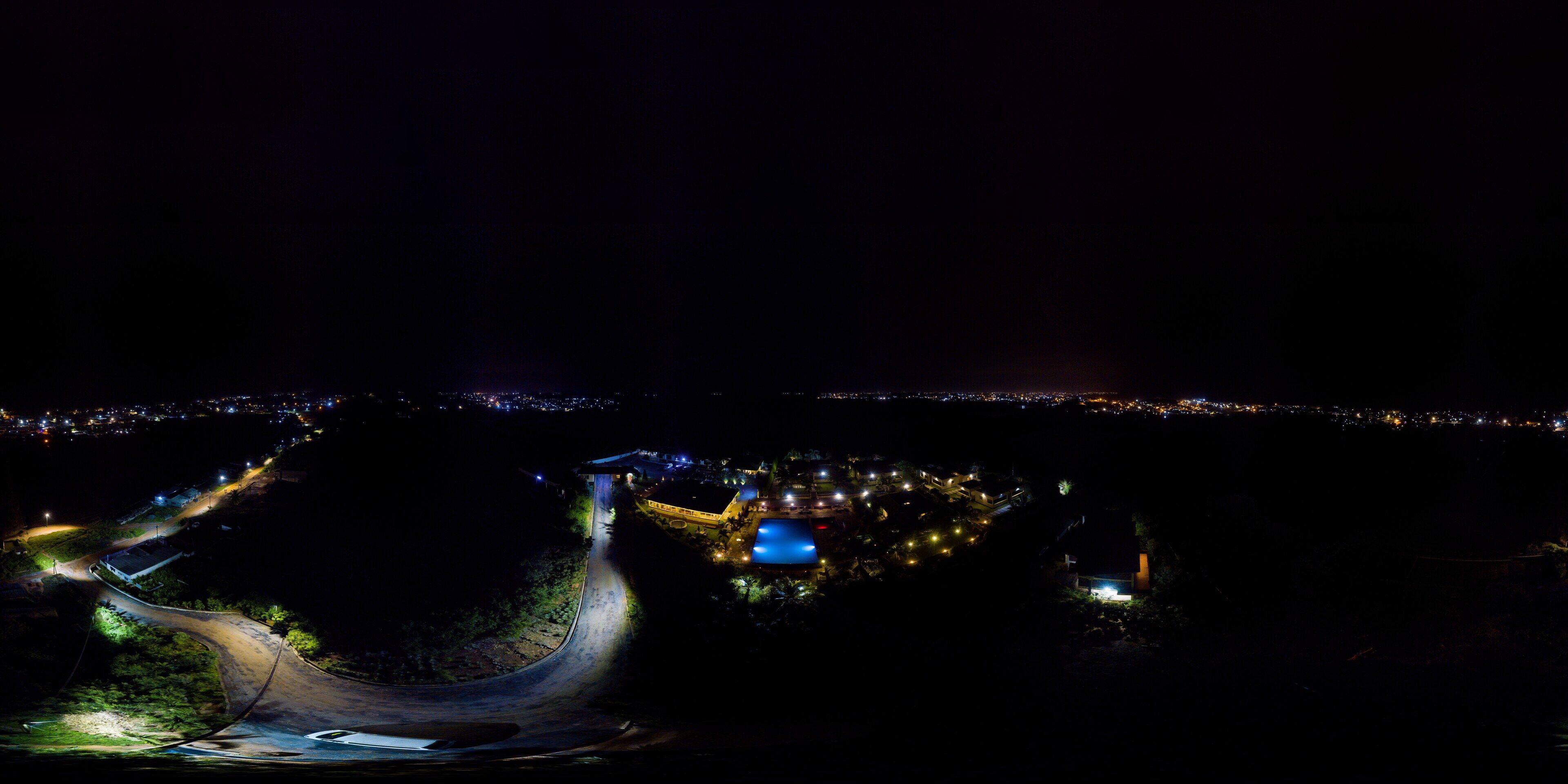 A nighttime aerial view of a winding road leading to a lit-up area with buildings and a bridge.