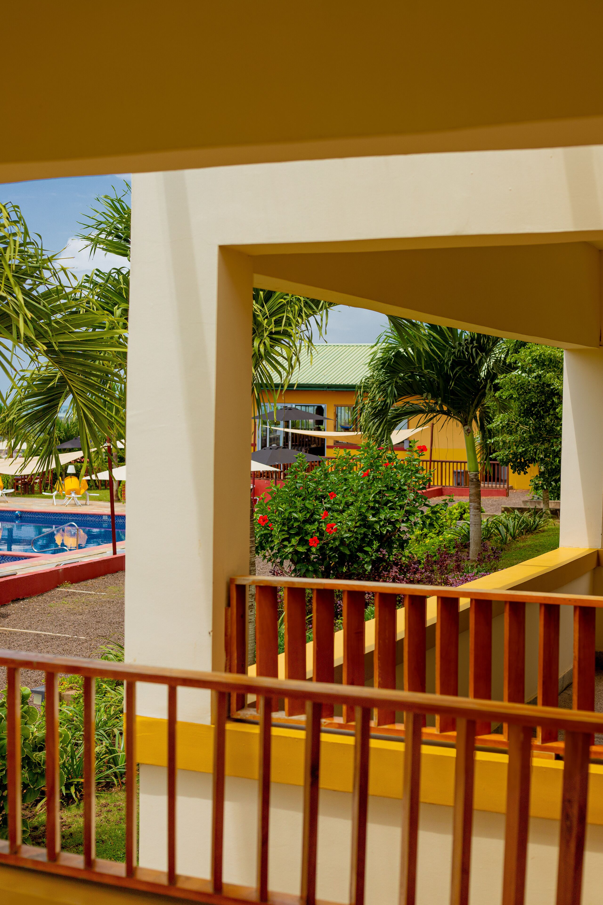 A view from a balcony with a pool, palm trees, and a building with yellow and red colors.