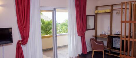 Habitación de hotel con puerta corrediza de vidrio al exterior, una cama, un escritorio con silla, un televisor y un estante de madera con un televisor y algunos objetos.