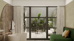 Une salle de bain moderne avec une baignoire sur pieds, un canapé vert et une vue sur un jardin tropical, visible à travers une grande porte vitrée.