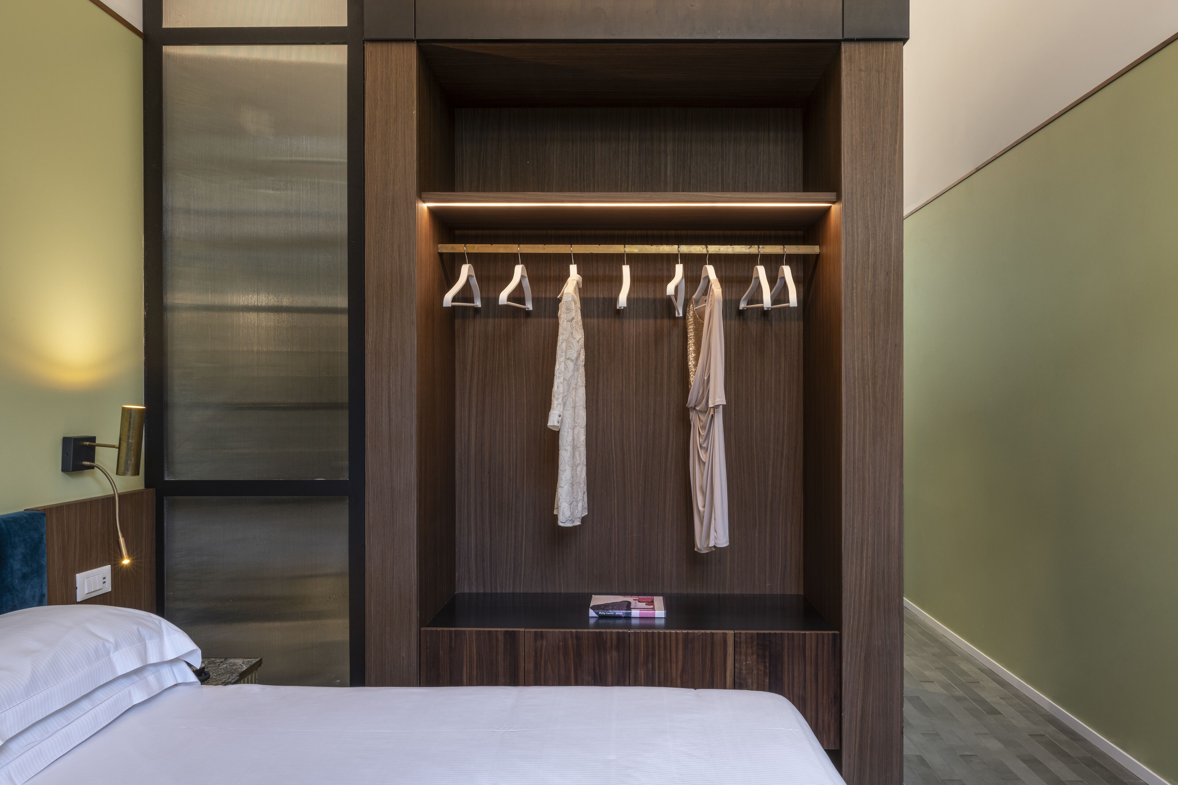Une chambre d’hôtel avec un lit, une armoire en bois avec une tringle pour suspendre les vêtements et une petite étagère avec un livre.