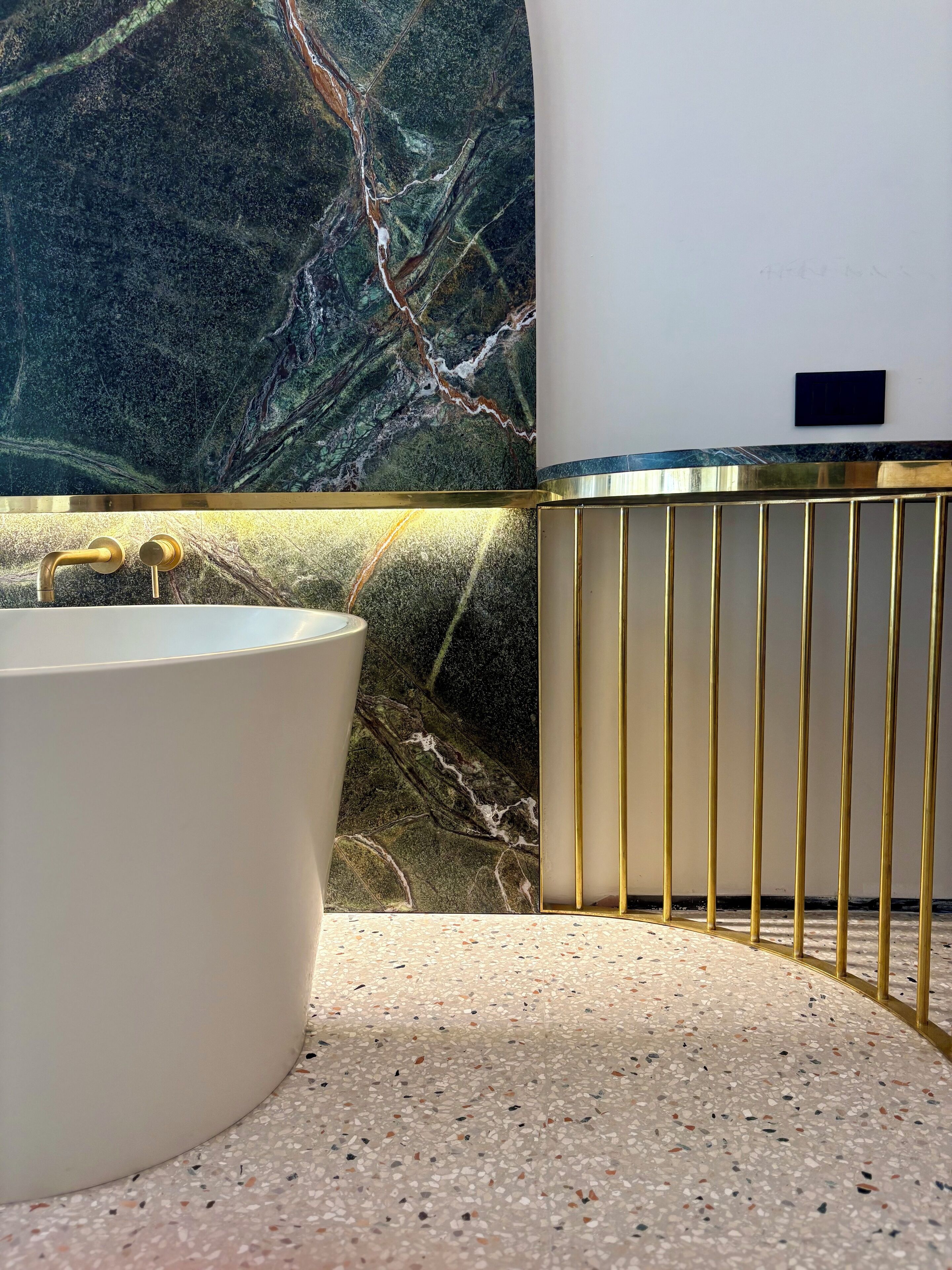 A bathroom with a marble wall, a white bathtub, and a gold-framed vanity.