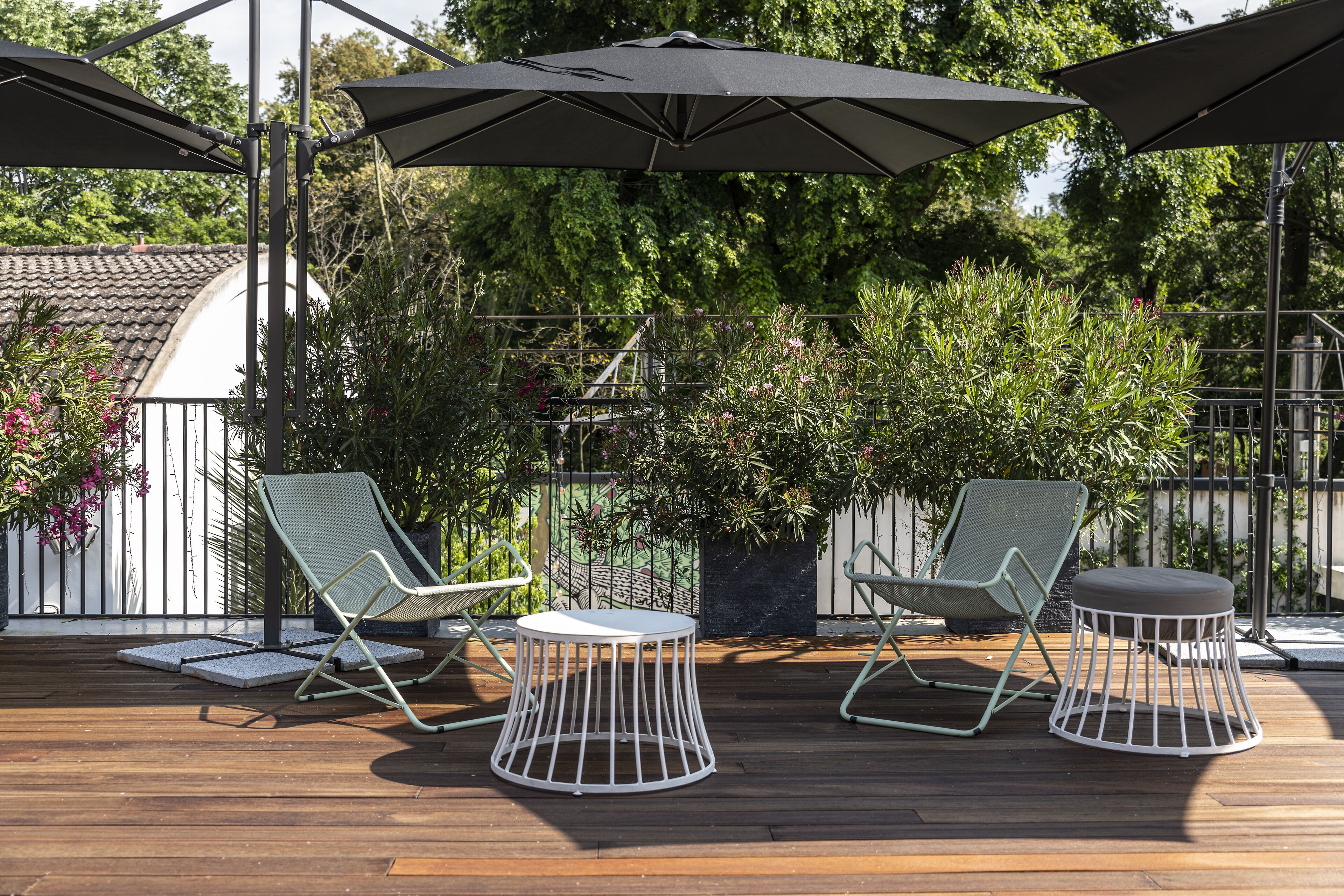 Una terraza de madera con dos sillones de descanso y una mesa redonda, rodeada de follaje y una valla de metal.