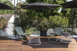 A wooden deck with two lounge chairs and a round table, surrounded by greenery and a metal fence.