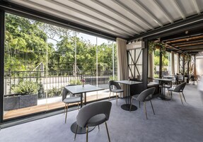 A modern outdoor dining area with tables and chairs, large windows offering a view of greenery, and a covered section with a wooden ceiling.