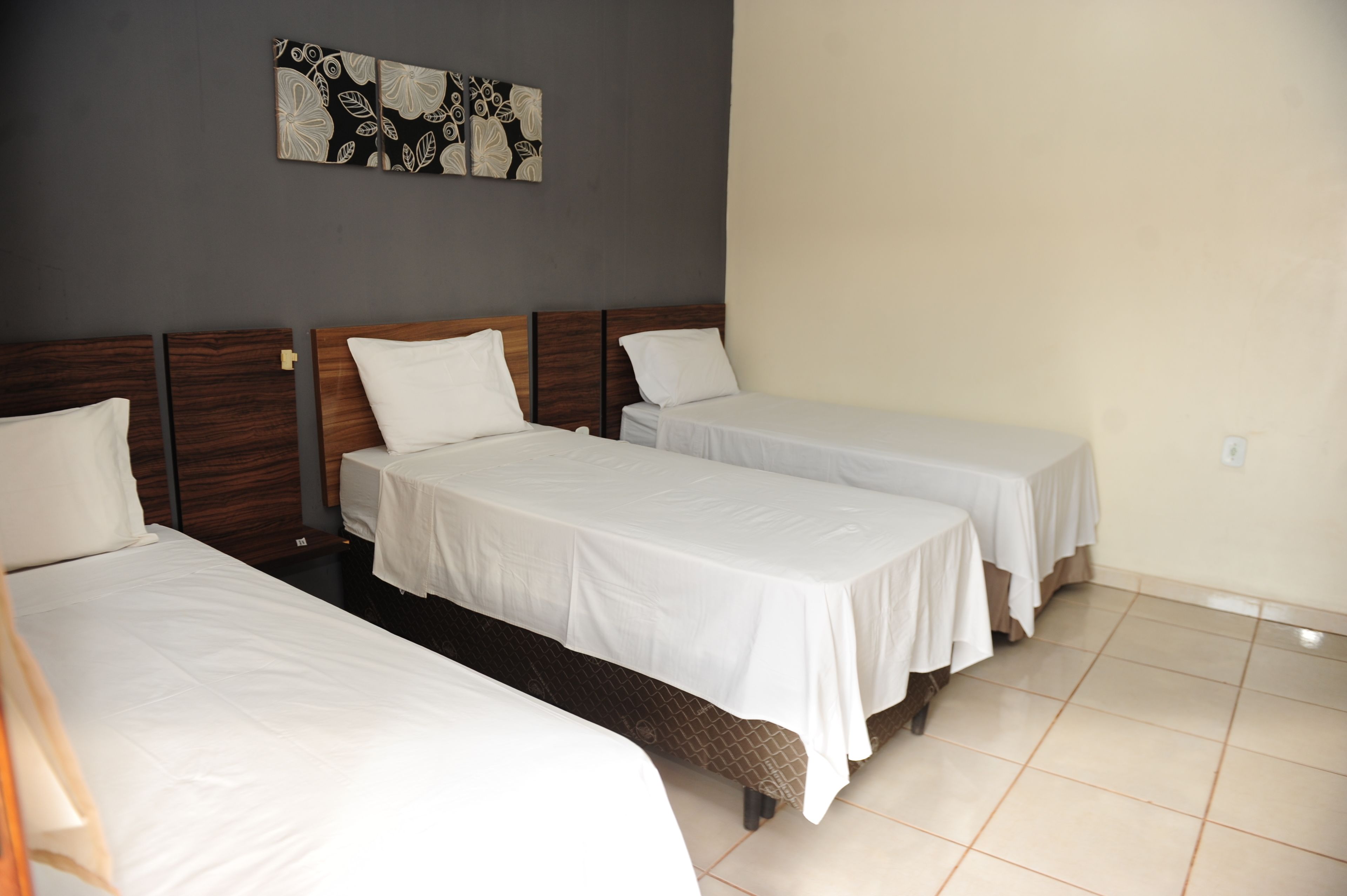 Habitación de hotel con tres camas individuales, un cabecero de madera oscura, piso de baldosas y una pared decorada con una obra de arte floral.