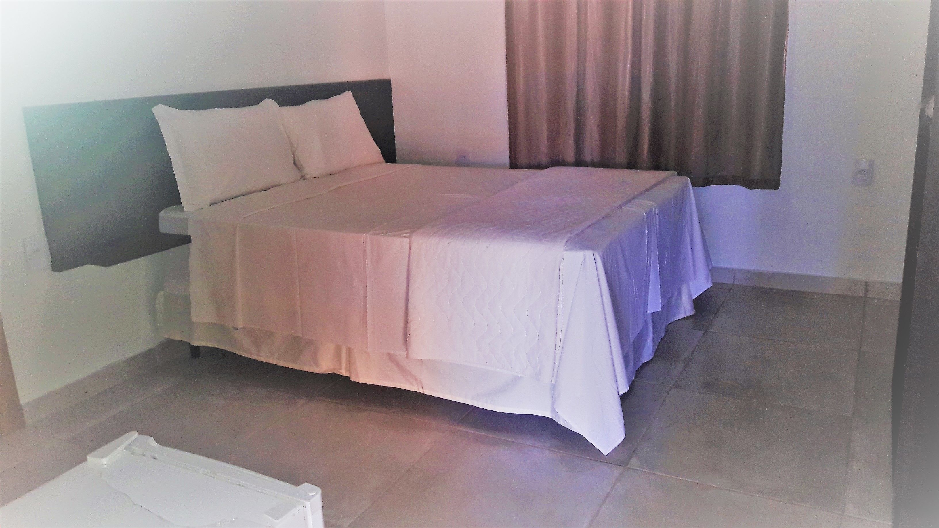 A neatly made bed with a white bedspread and pillows, a bedside table with a lamp, and a window with curtains.