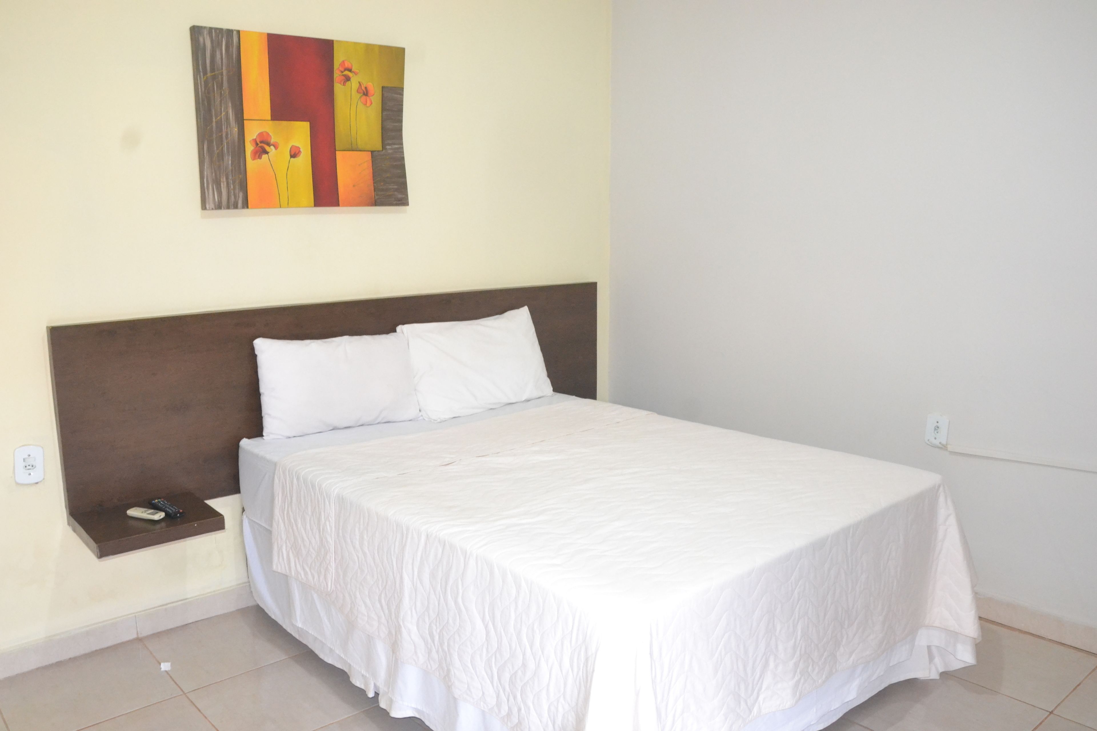 A neatly made bed with a wooden headboard, a bedside table with a remote control, and a wall-mounted artwork.