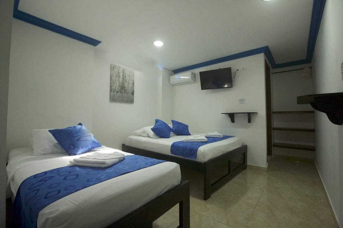 A hotel room with two beds, each with blue bedding and pillows, a flat-screen TV mounted on the wall, and an air conditioning unit.