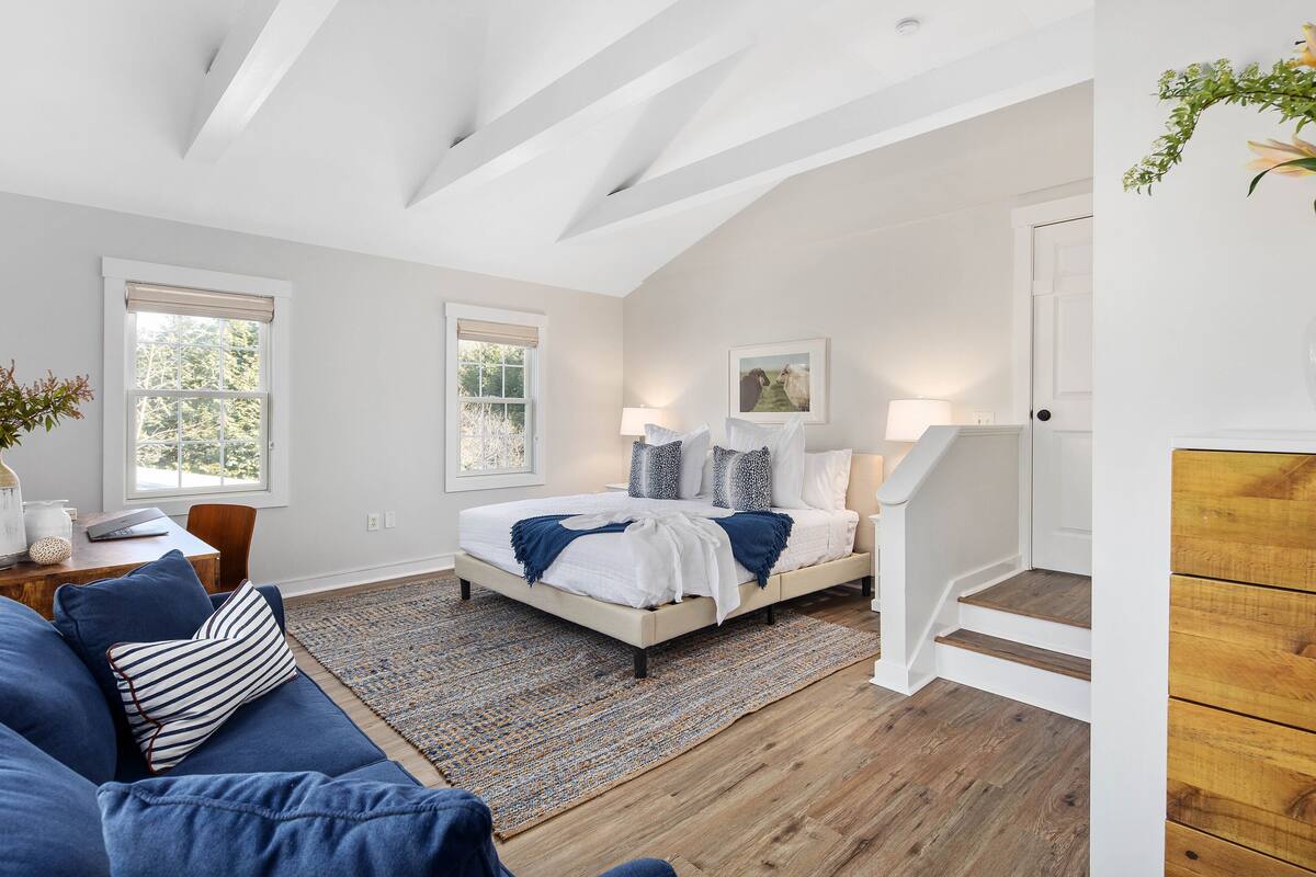 A bedroom with a bed, a blue sofa, a wooden staircase, and a view of the outdoors.