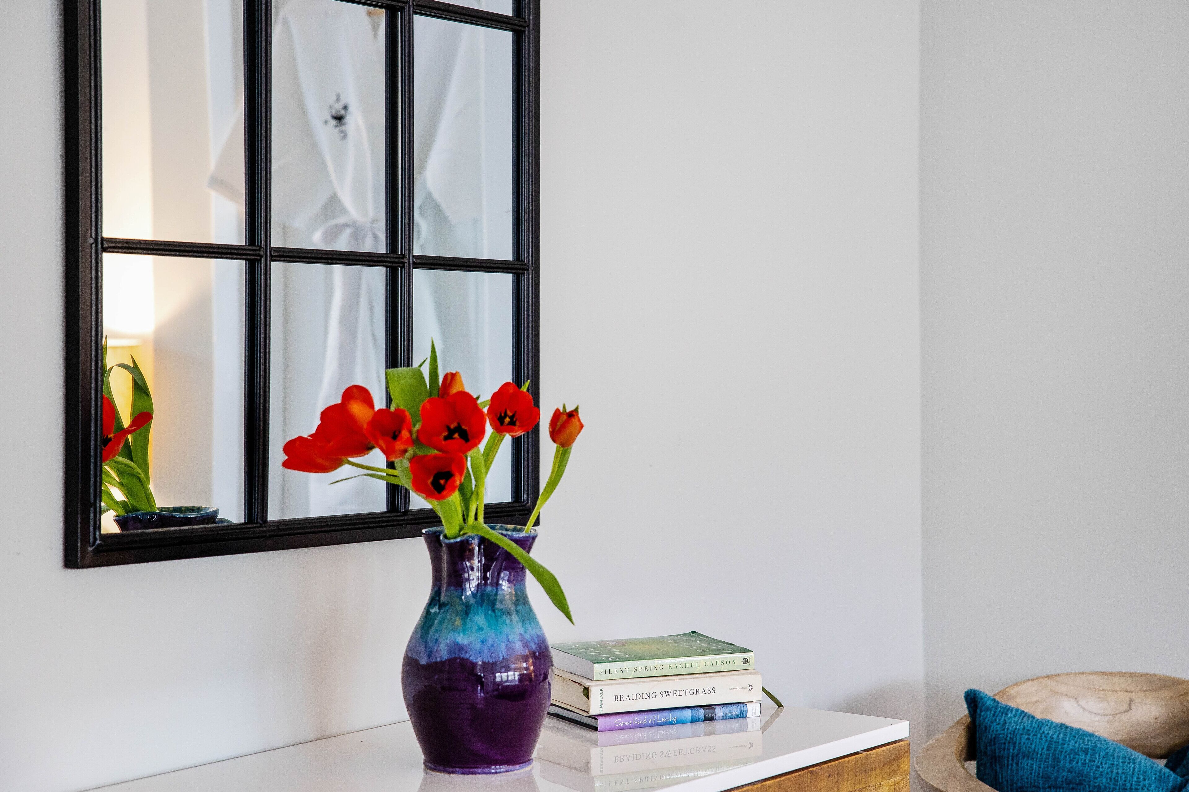 A purple vase with red flowers on a white surface next to a window with a white curtain.