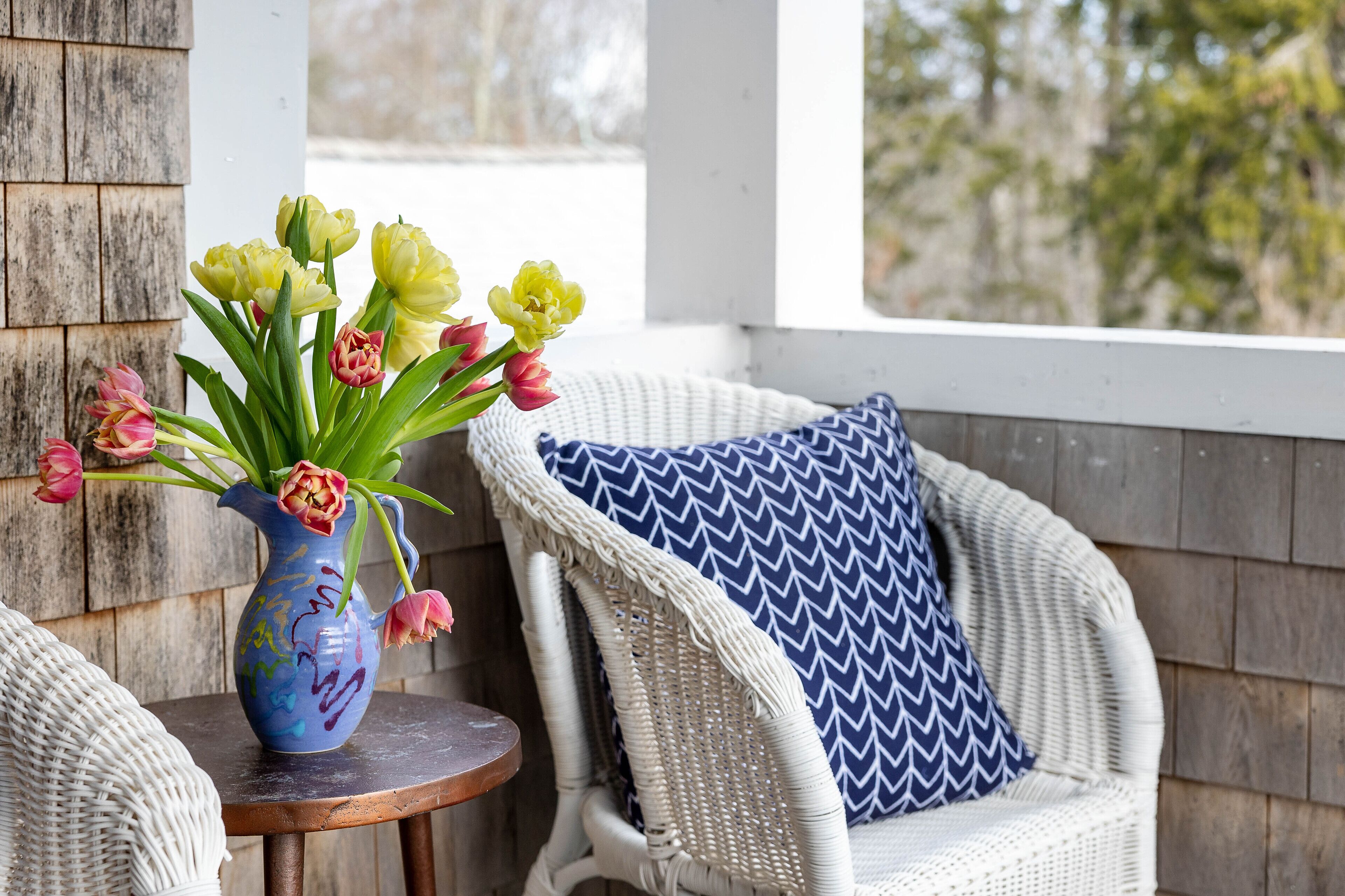 Una silla de mimbre con un cojín de motivos azules y blancos, una mesita redonda con un jarrón de flores y una pared de madera.