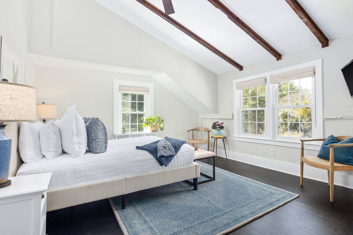 A bedroom with a bed, bedside table, lamp, windows, and a chair.