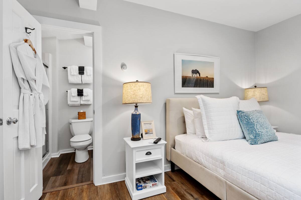 A hotel room with a bed, bedside lamps, a nightstand, a framed picture on the wall, and a bathroom visible through an open door.