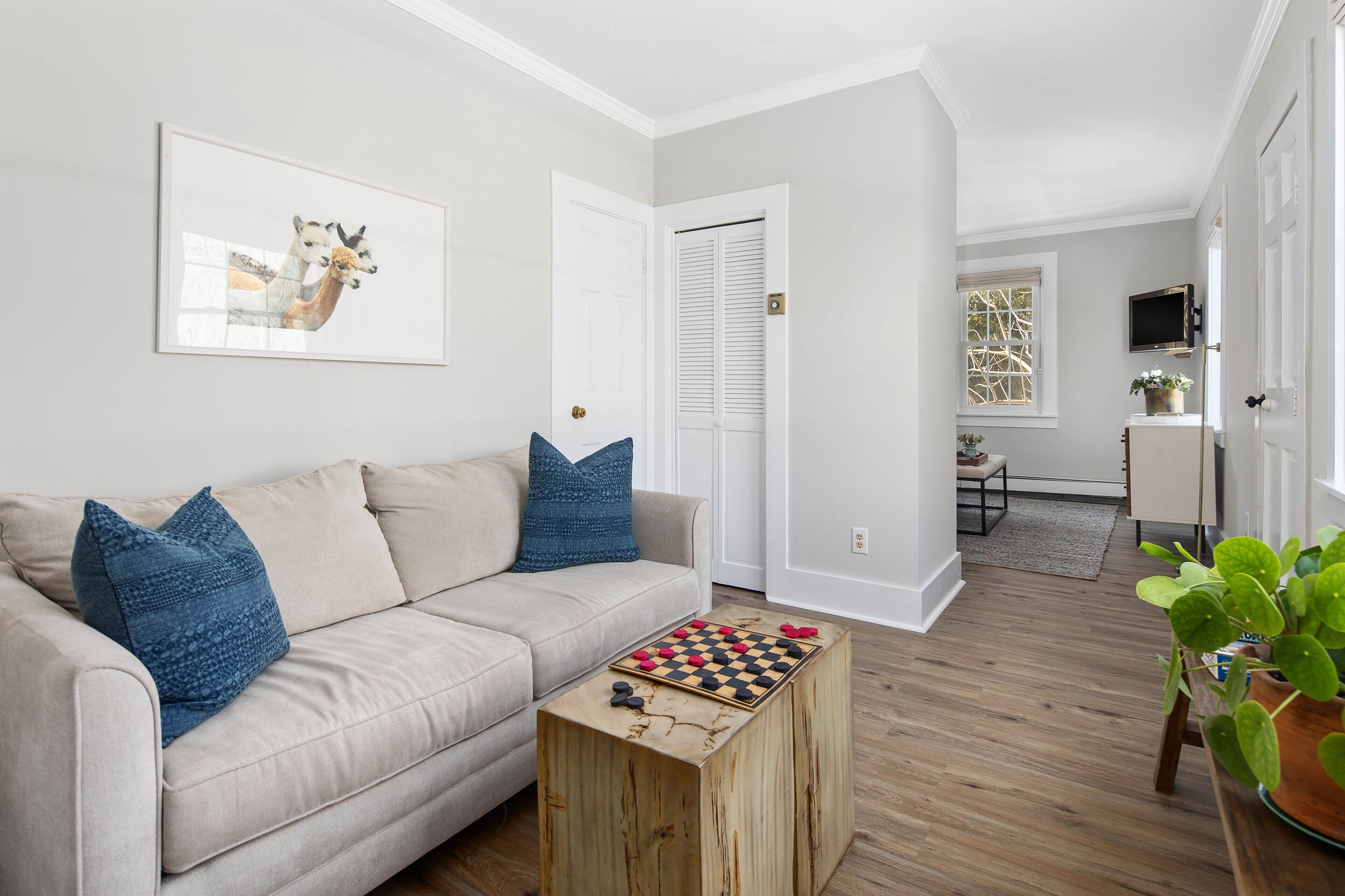 Una sala de estar con un sofá beige, almohadas azules, una mesa de centro de madera con un tablero de ajedrez y un cuadro en la pared.