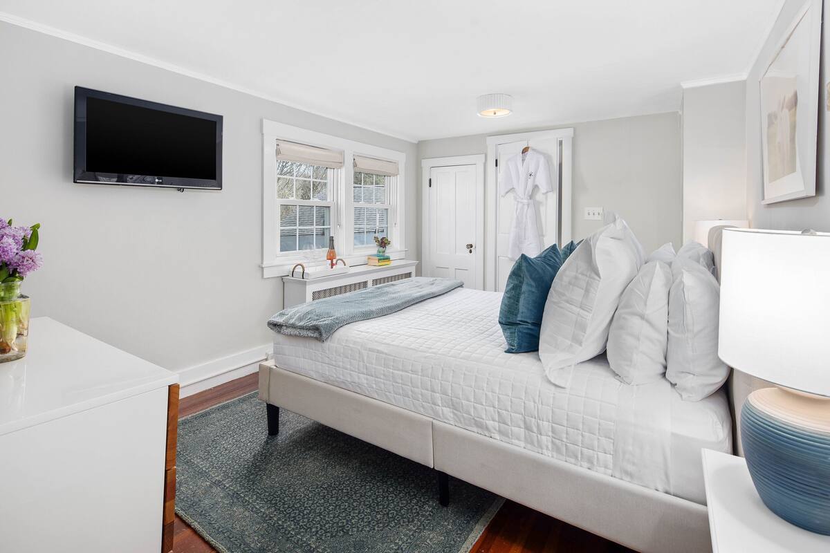 A bedroom with a bed, a flat-screen TV, a window with blinds, a white door, and a robe hanging on the door.