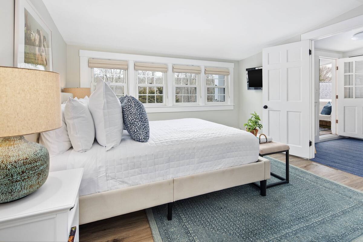 A bedroom with a bed, a nightstand, a lamp, a window with blinds, and a door.