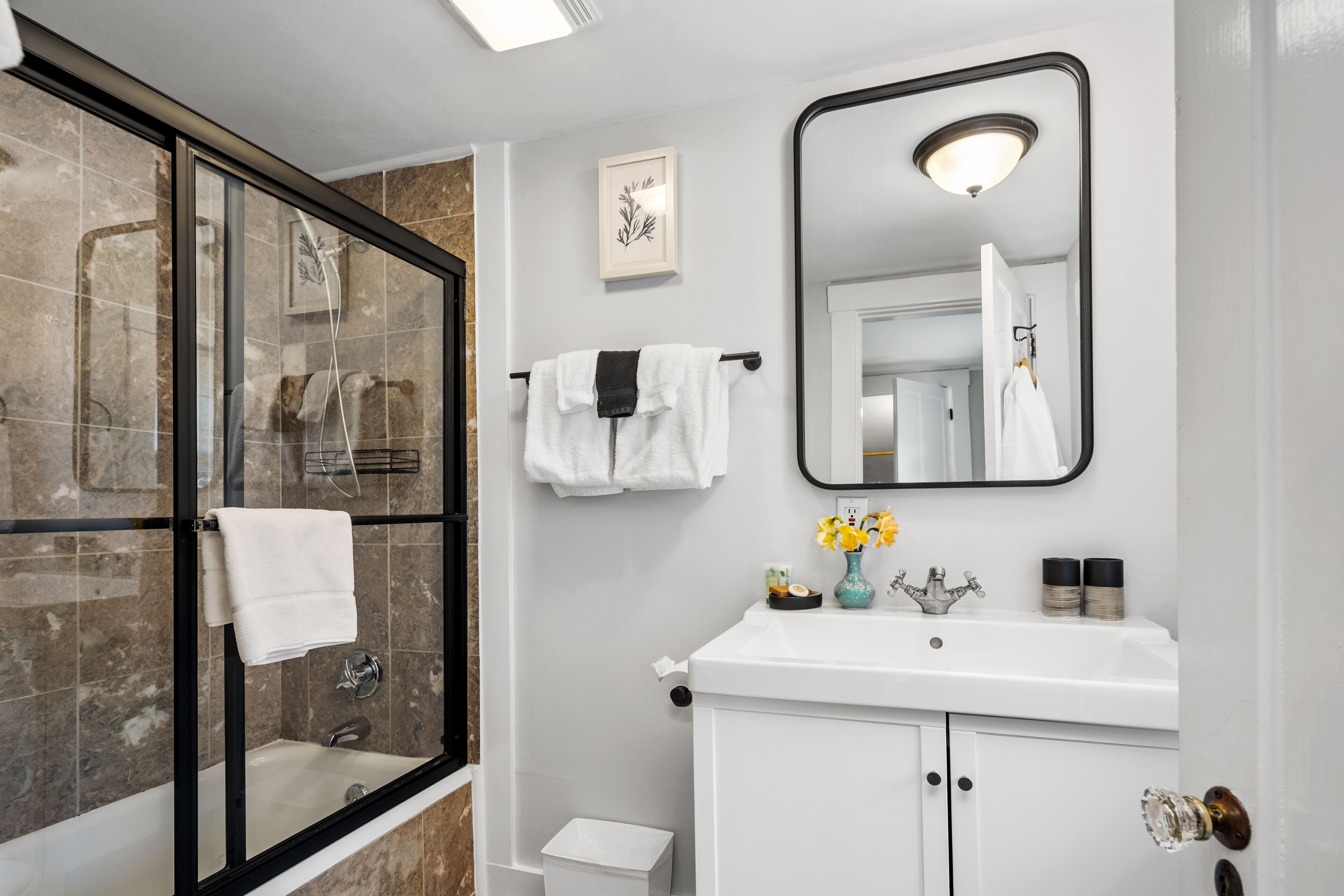 A modern bathroom with a glass-enclosed shower, a white sink with a vase of flowers, and a large mirror.