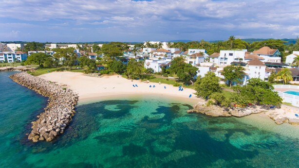 A coastal resort with clear turquoise waters, a sandy beach, and a row of white buildings with blue roofs. - Point Village Studios and Suites (Negril)