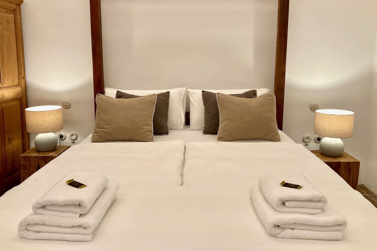 A neatly made bed with white linens and two folded towels on each side, flanked by two bedside lamps and a wooden headboard.