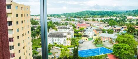 A residential area with a tennis court, a multi-story building, and a view of a hilly landscape.