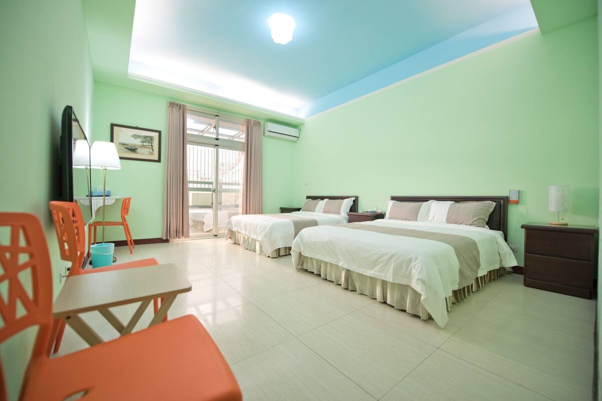 A hotel room with two beds, a desk, chairs, a TV, and a window with curtains.