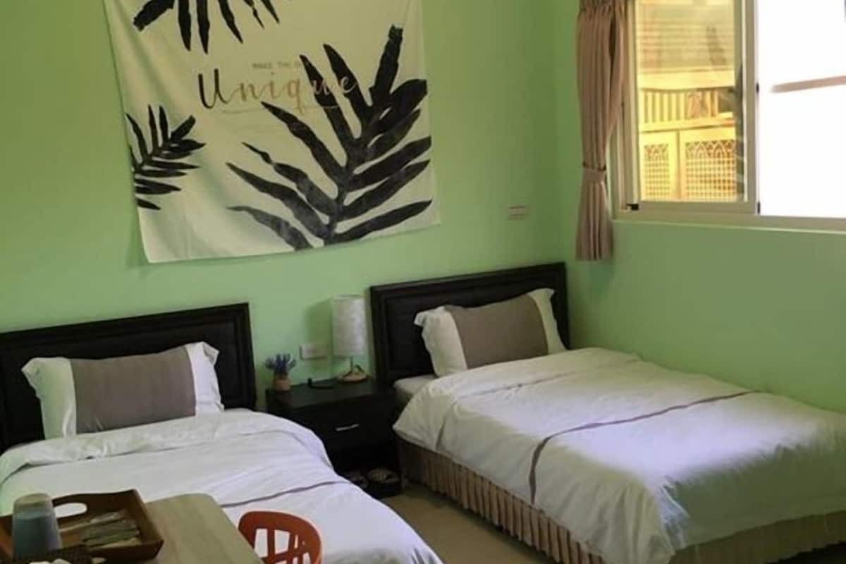 A room with two beds, a desk with a chair, a large wall tapestry, and a window with curtains.