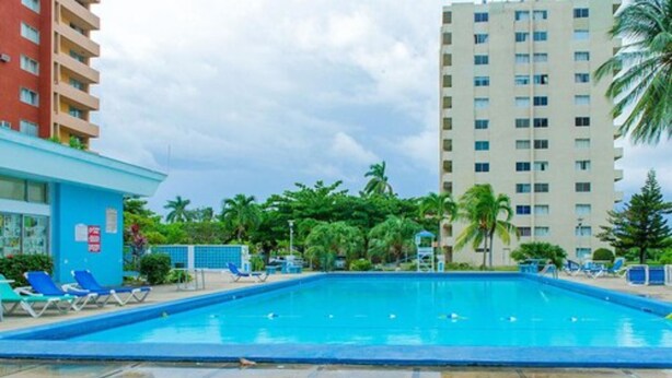 A swimming pool with lounge chairs, surrounded by palm trees and a tall building. - Sword Fish Beach Suite at Turtle Towers (Ocho Rios)