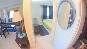 A compact hotel room with a bed, a desk, a chair, a lamp, and a round mirror. - Sword Fish Beach Suite at Turtle Towers (Ocho Rios)