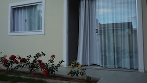 Un bâtiment doté de deux fenêtres : l’une est fermée et munie d’un store blanc, l’autre est ouverte et présente une porte coulissante. Devant le bâtiment, on peut voir des fleurs rouges et jaunes.