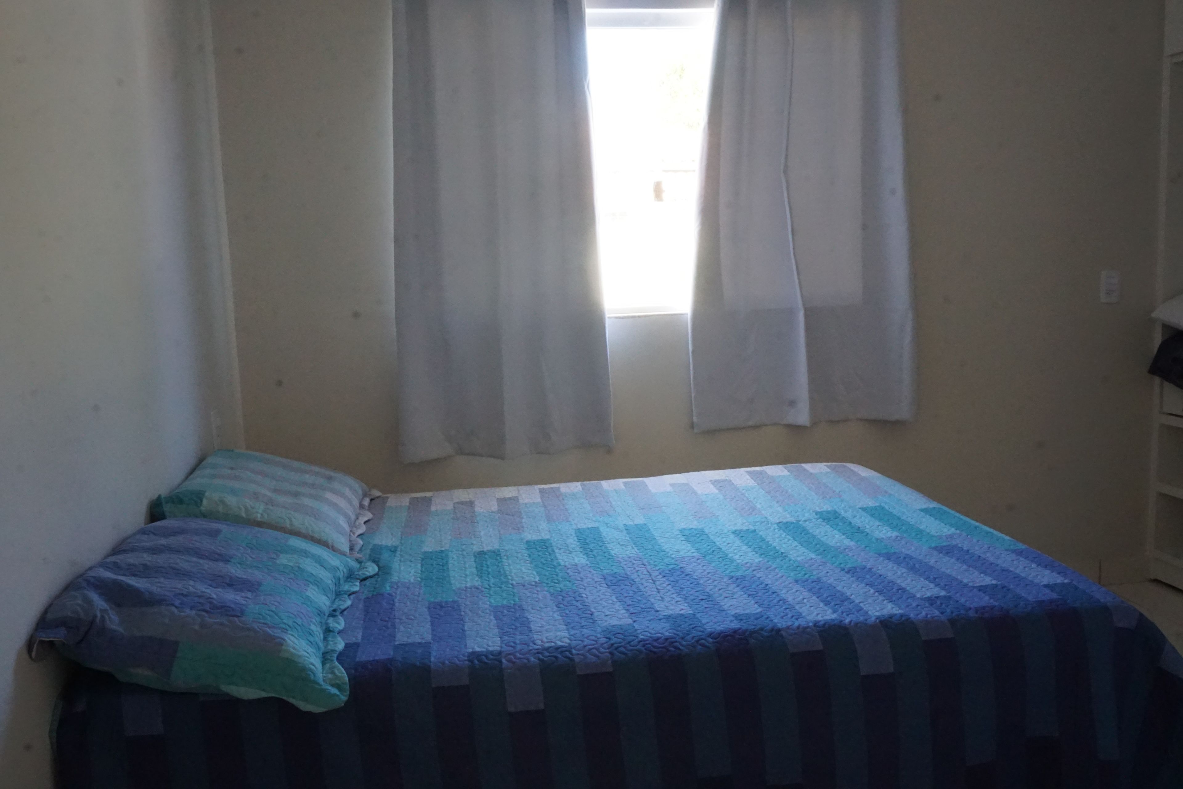 Una habitación pequeña con cama individual, una colcha estampada, un almohadón y cortinas de encaje.
