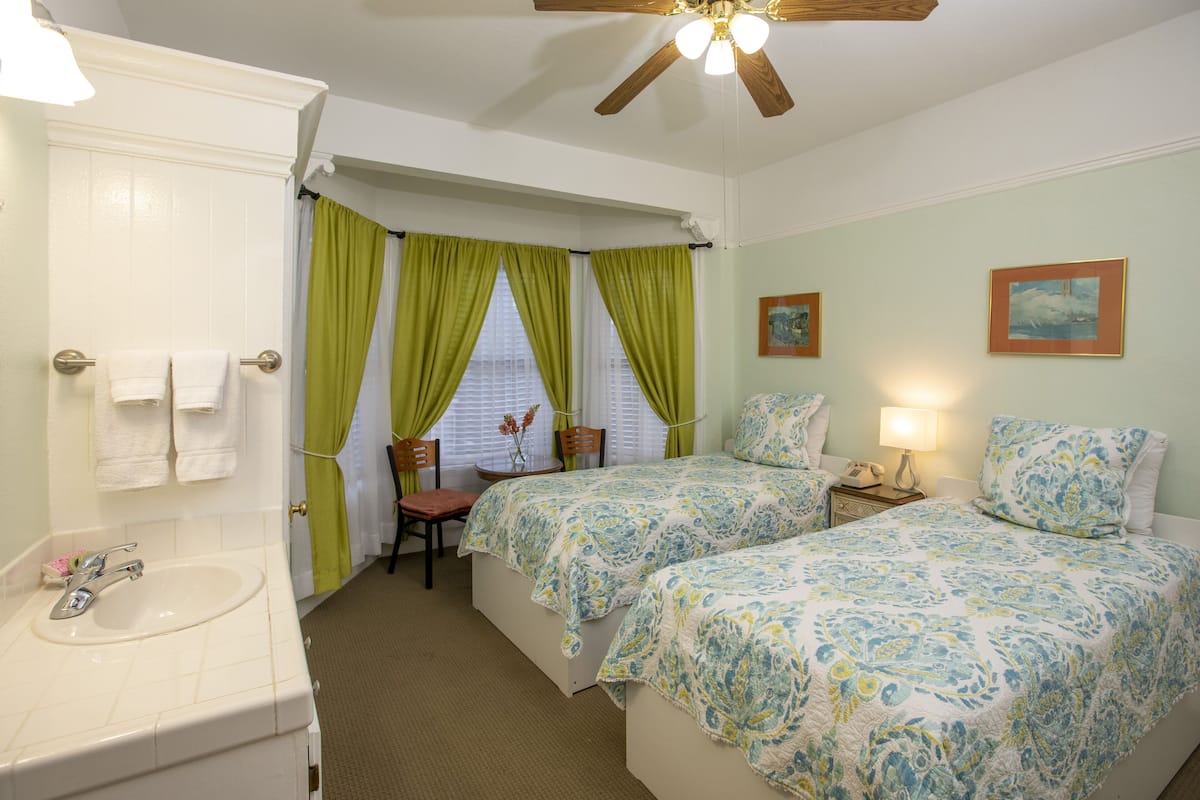A hotel room with a bathroom, two beds with floral bedding, a ceiling fan, and a window with green curtains.