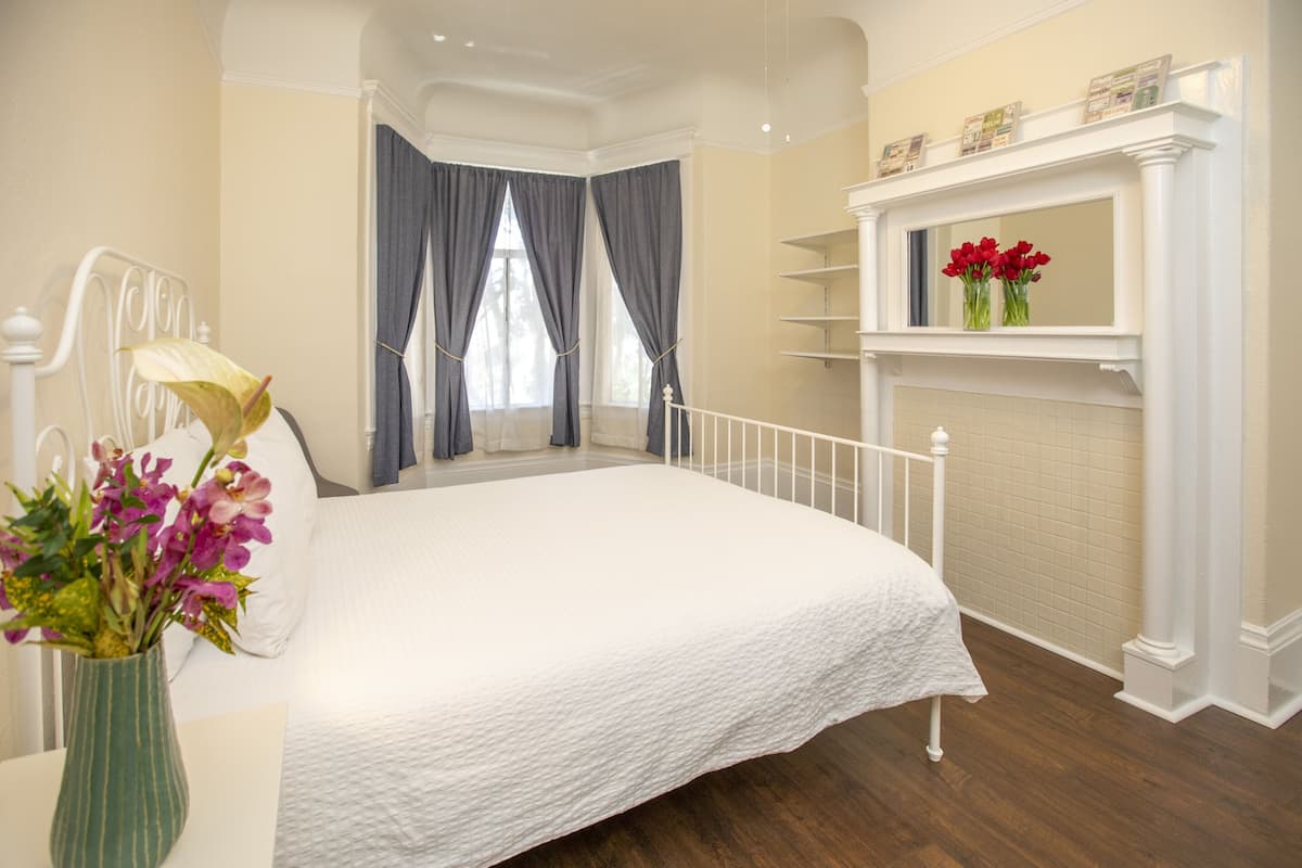 A bedroom with a large bed, a white headboard, a vase with flowers, a fireplace with a mantel, and a window with curtains.