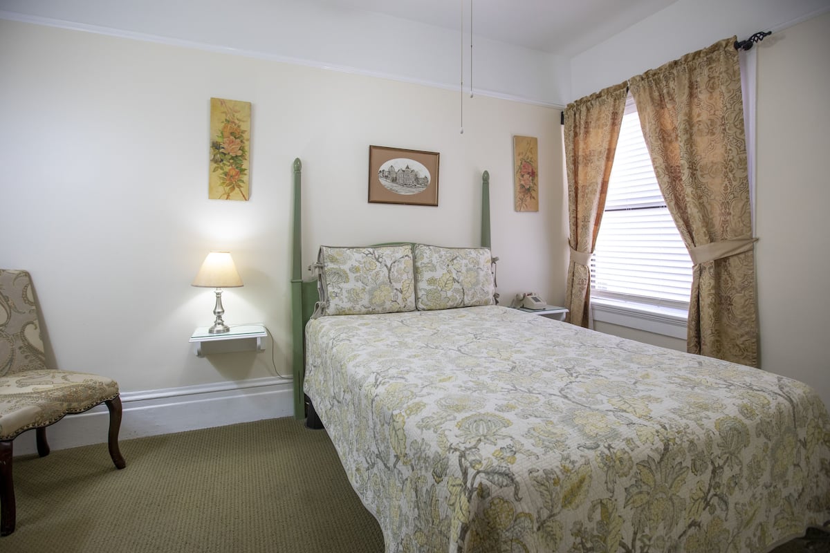 A bedroom with a bed, a chair, a nightstand with a lamp, and two wall-mounted paintings.