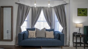 A living room with a blue sofa, grey curtains, a mirror, and a small table with a lamp. - Hayes Valley Inn (San Francisco)