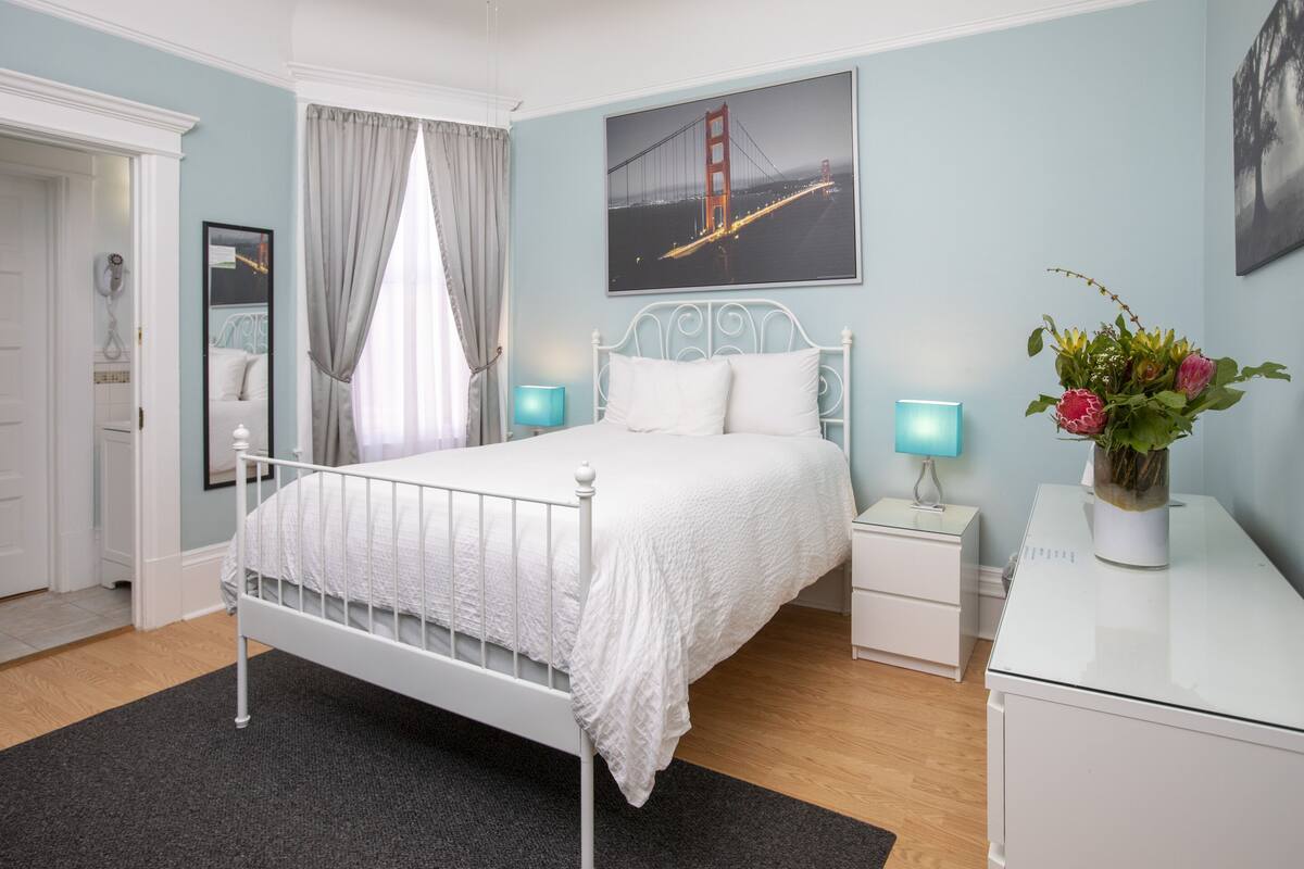A bedroom with a white metal bed, a nightstand with a lamp, a dresser, and a framed picture of the Golden Gate Bridge.