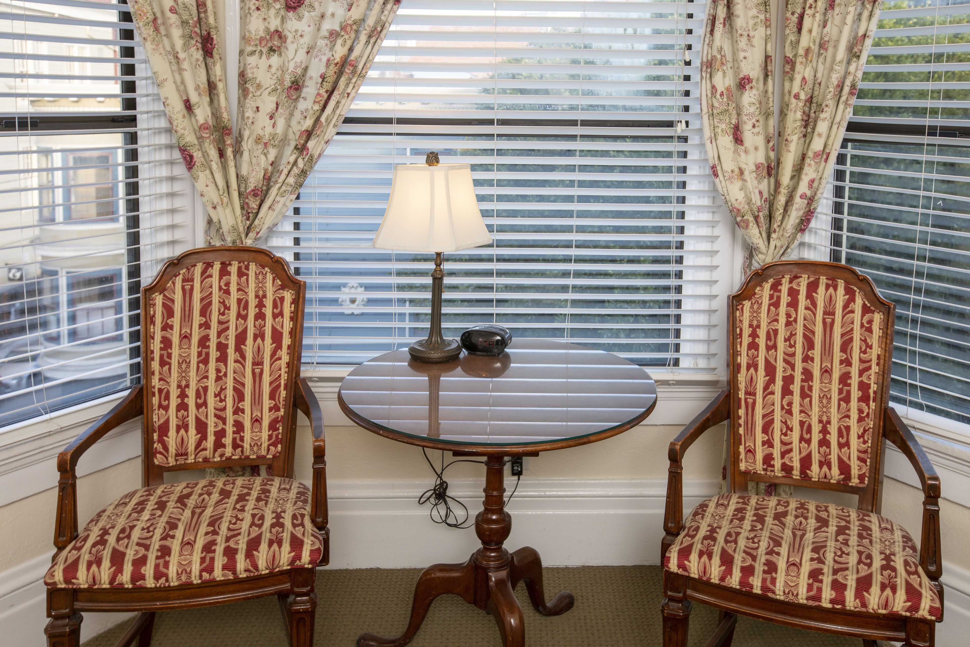 A corner room with two upholstered chairs, a small round table with a lamp, and a window with blinds and floral curtains.